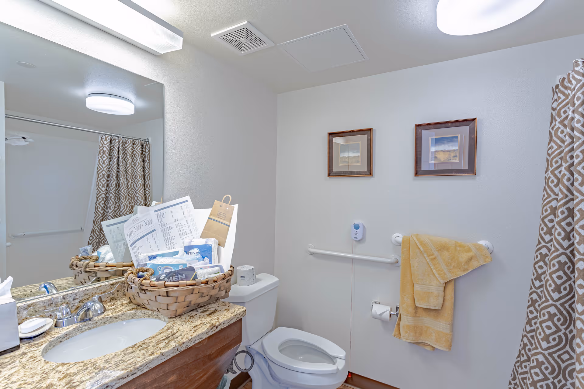 A clean and well-lit bathroom with a granite countertop, a sink, and a large mirror. There is a basket on the counter containing various items including papers and packages. The toilet is next to the counter with a toilet paper roll on the holder and a grab bar on the wall. Two framed pictures hang above the grab bar, and a yellow towel is draped over another grab bar. A patterned shower curtain is visible in the background.