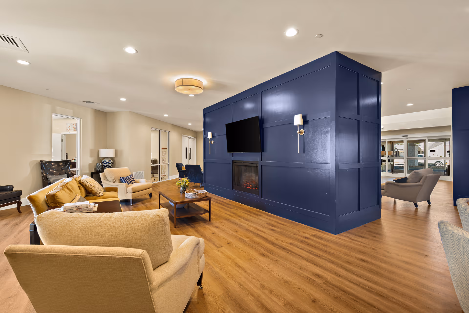 A spacious and well-lit living room area with wooden flooring, beige and blue armchairs, a beige sofa, and a wooden coffee table. A large blue paneled wall features a mounted flat-screen TV and a built-in electric fireplace. The room has recessed lighting and a ceiling light fixture, with large windows and glass doors visible in the background.