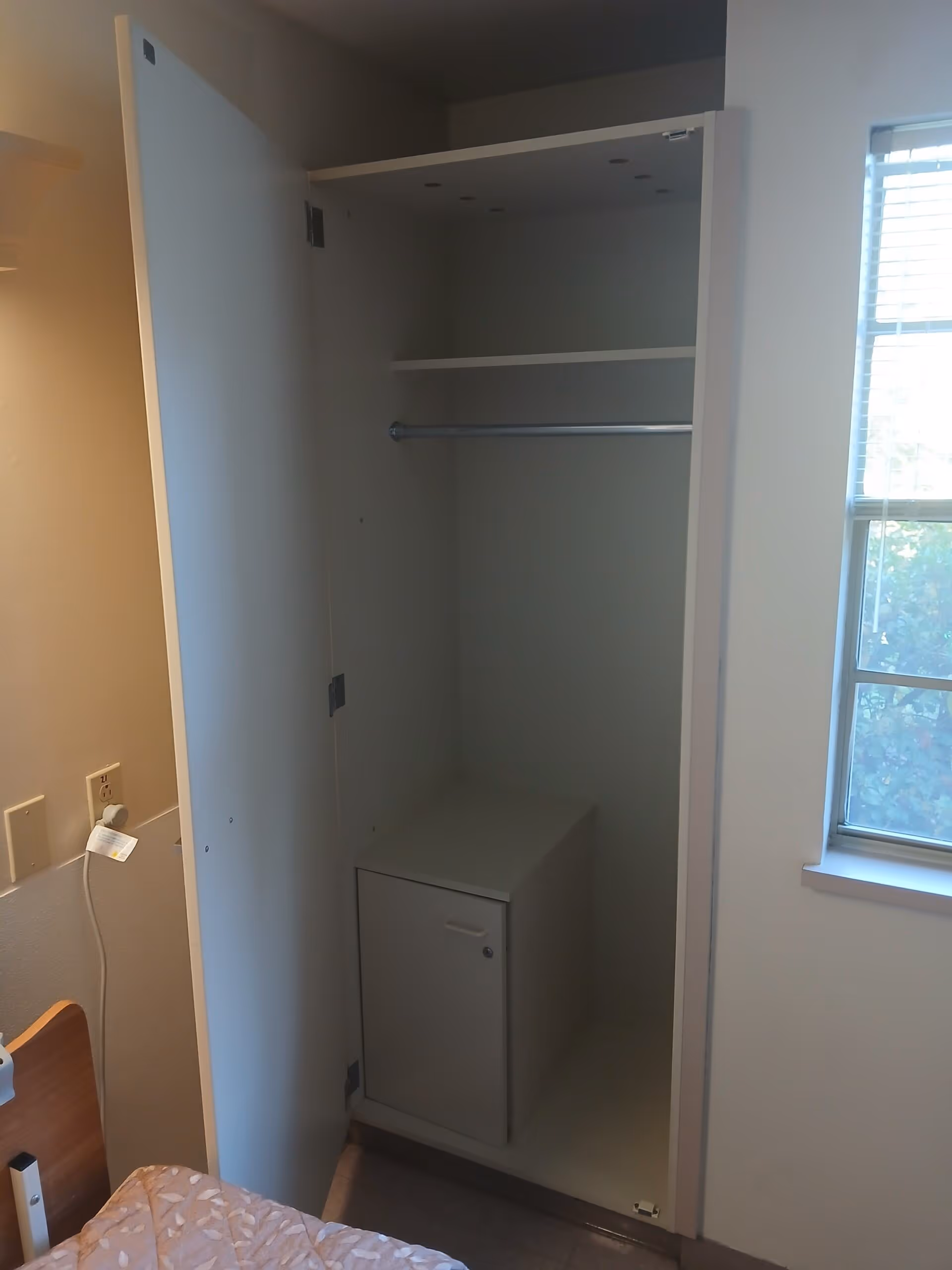 An open white closet inside a room with a hanging rod, a shelf, and a small cabinet at the bottom. To the right of the closet is a window with blinds partially open, and to the left is part of a bed and a wall outlet with a plugged-in device.