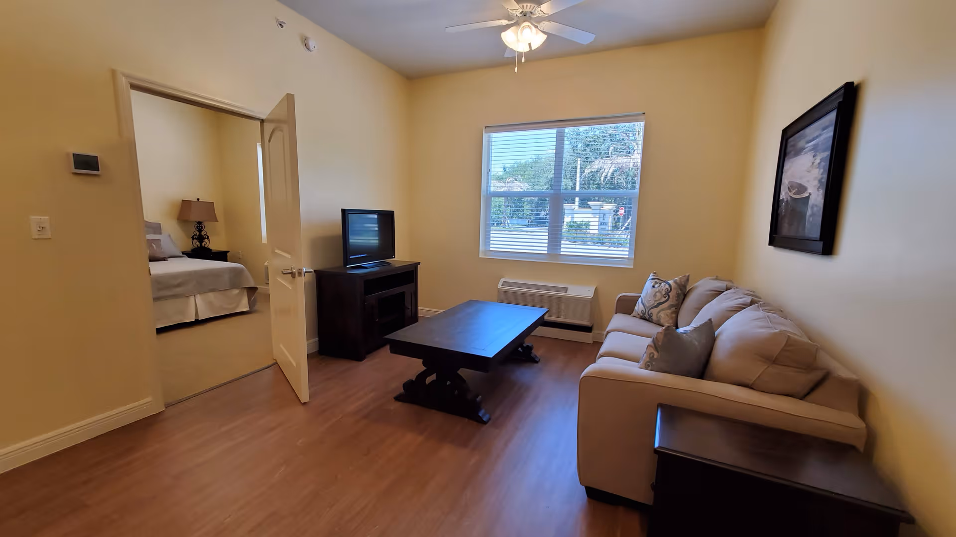 Small living room with a couch, coffee table, TV on a stand, window, and an open door leading to a bedroom.