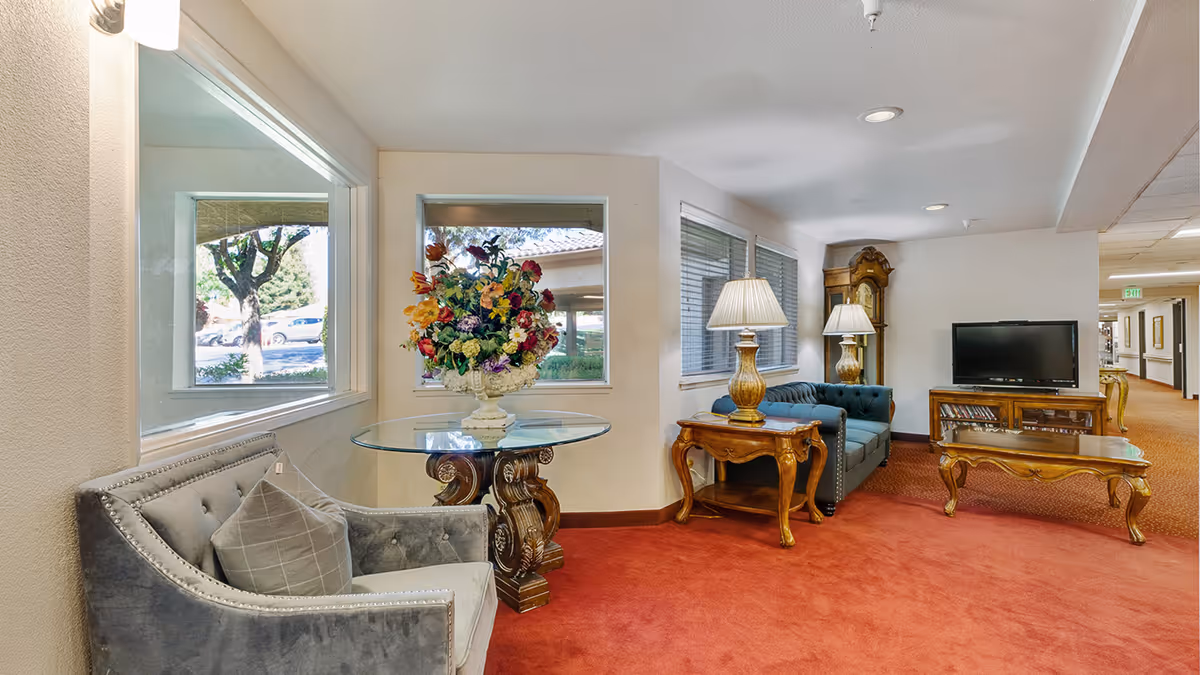 A cozy senior living community lounge area with a red carpet, a gray armchair with a cushion, a glass-top table with a large floral arrangement, a wooden side table with a lamp, a blue tufted sofa, a wooden coffee table, a wooden TV stand with a flat-screen TV, and a grandfather clock. Windows provide natural light and a view of the outside.