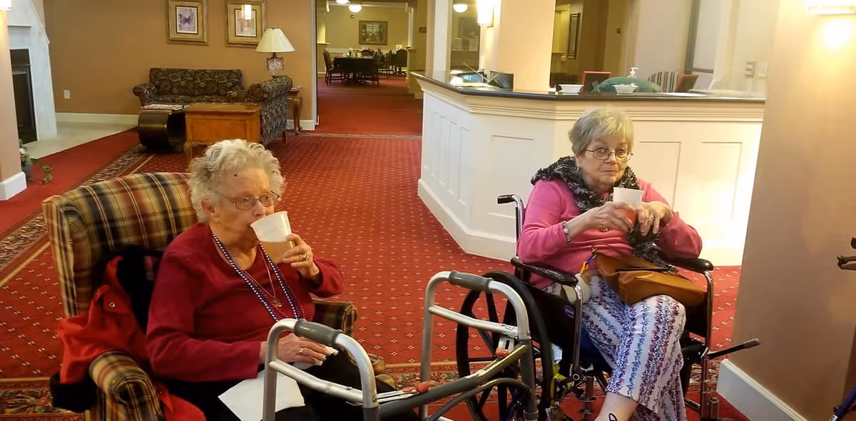 Two elderly women seated in a senior living common room, one in an armchair and one in a wheelchair, drinking from cups.