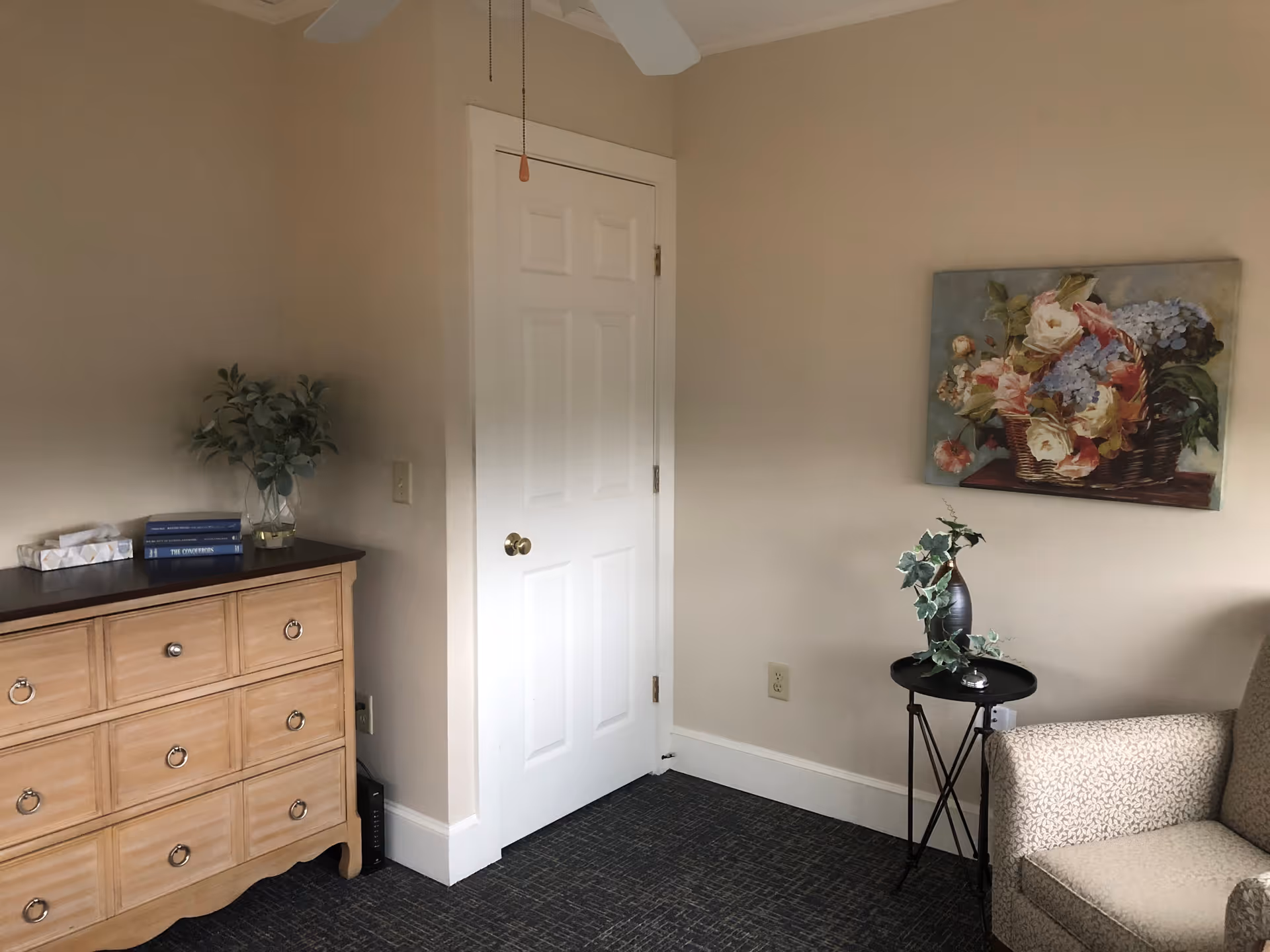A corner of a room with a wooden dresser topped with a tissue box, two books, and a glass vase with green foliage. A white door with a brass knob is centered on the wall. To the right, there is a small black round table with a vase holding green ivy and a silver bell. A beige patterned armchair is partially visible. A painting of a basket of flowers hangs on the wall above the table. The room has beige walls, a dark carpeted floor, and a white ceiling fan pull chain hanging down.