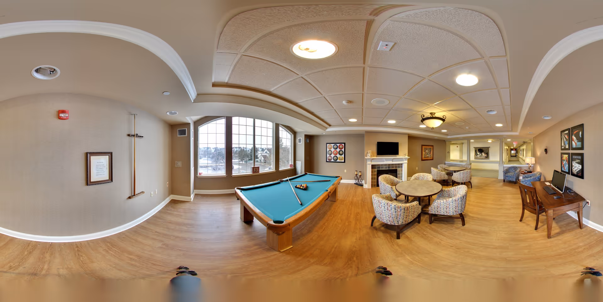A spacious senior living common area featuring a pool table in the center, a round table with four upholstered chairs, a fireplace with a mounted TV above it, a computer desk with two monitors, and large windows letting in natural light. The room has wooden flooring, beige walls, and ceiling lights, with framed artwork on the walls.