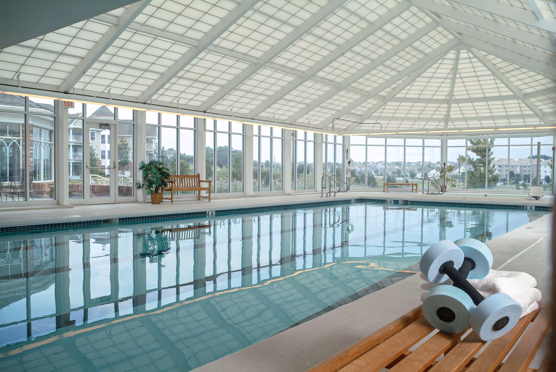 Indoor swimming pool with large windows and a glass ceiling allowing natural light. Wooden benches and potted plants are placed around the pool area. A pair of light blue dumbbells and white towels are on a wooden bench in the foreground.
