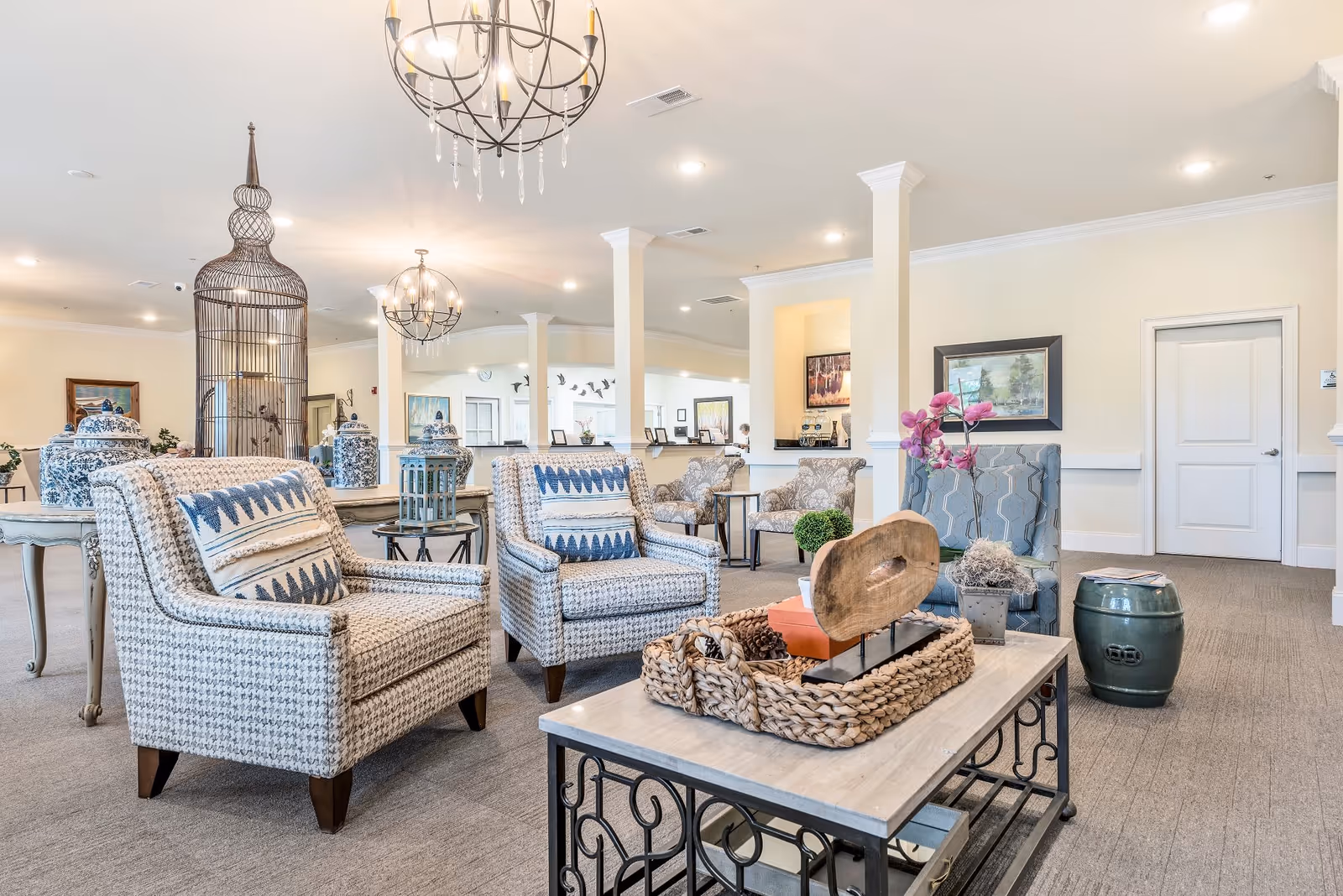 A bright and spacious senior living facility common area with patterned armchairs, a coffee table decorated with a woven basket and ornamental items, chandeliers hanging from the ceiling, and decorative vases and artwork on the walls.