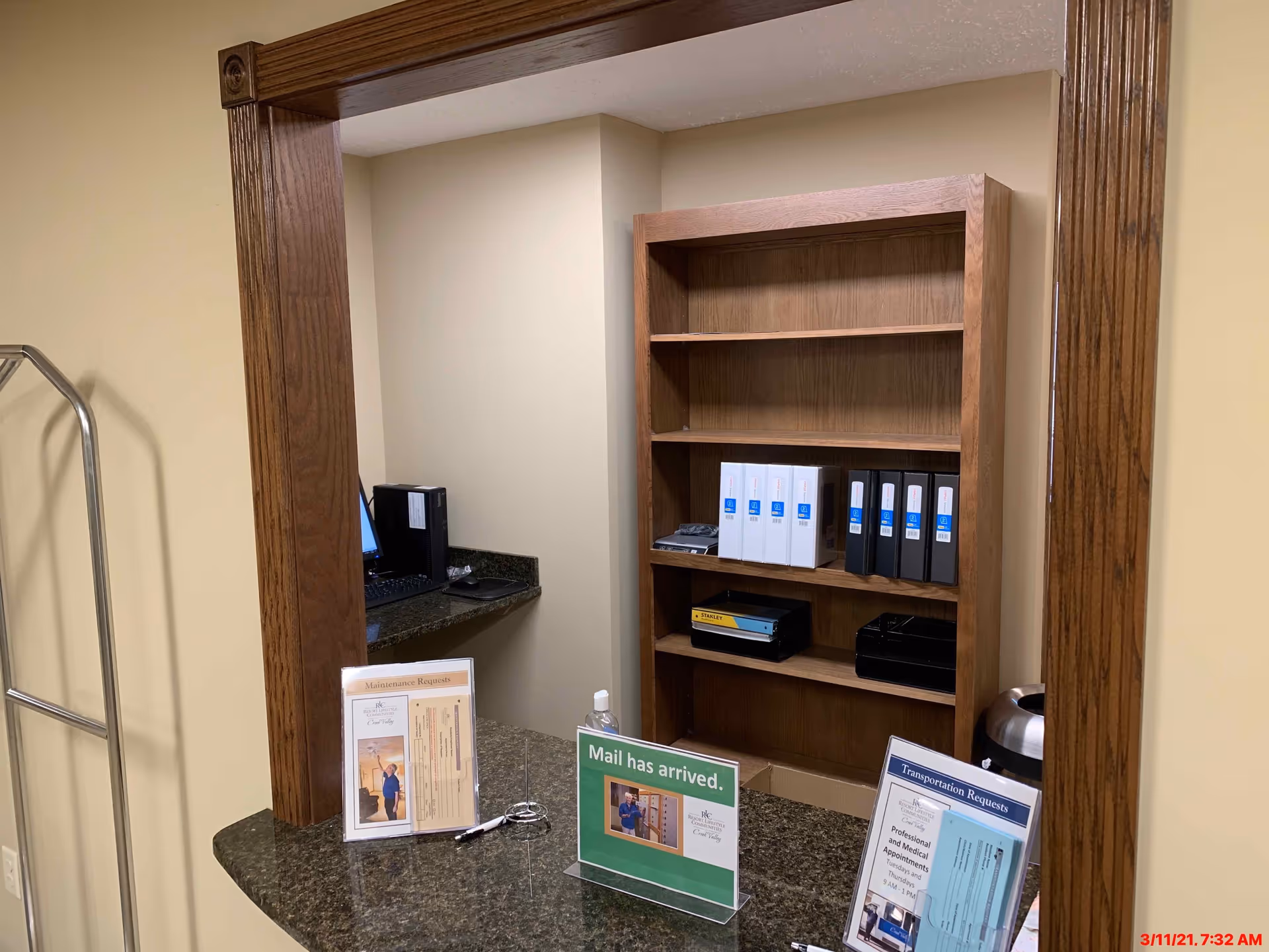 Reception or front desk area with a wooden framed window opening. Behind the desk is a wooden bookshelf with binders and office supplies. On the counter are informational signs about maintenance requests, mail arrival, and transportation requests. A computer monitor and keyboard are partially visible in the background.