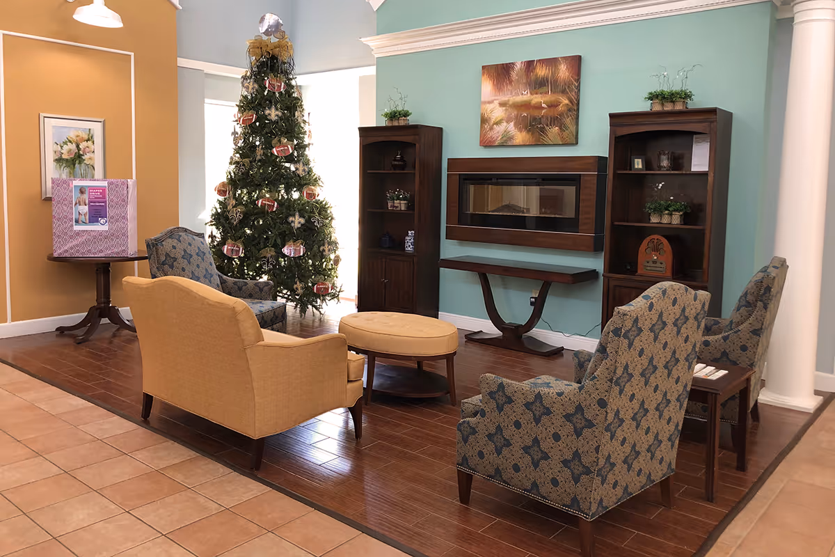 A cozy living room area with a Christmas tree decorated with football ornaments, two patterned armchairs, a yellow loveseat, a round ottoman, and two wooden bookshelves flanking a modern electric fireplace mounted on a teal accent wall. A framed painting hangs above the fireplace, and a small table with a pink donation box is visible on the left side.