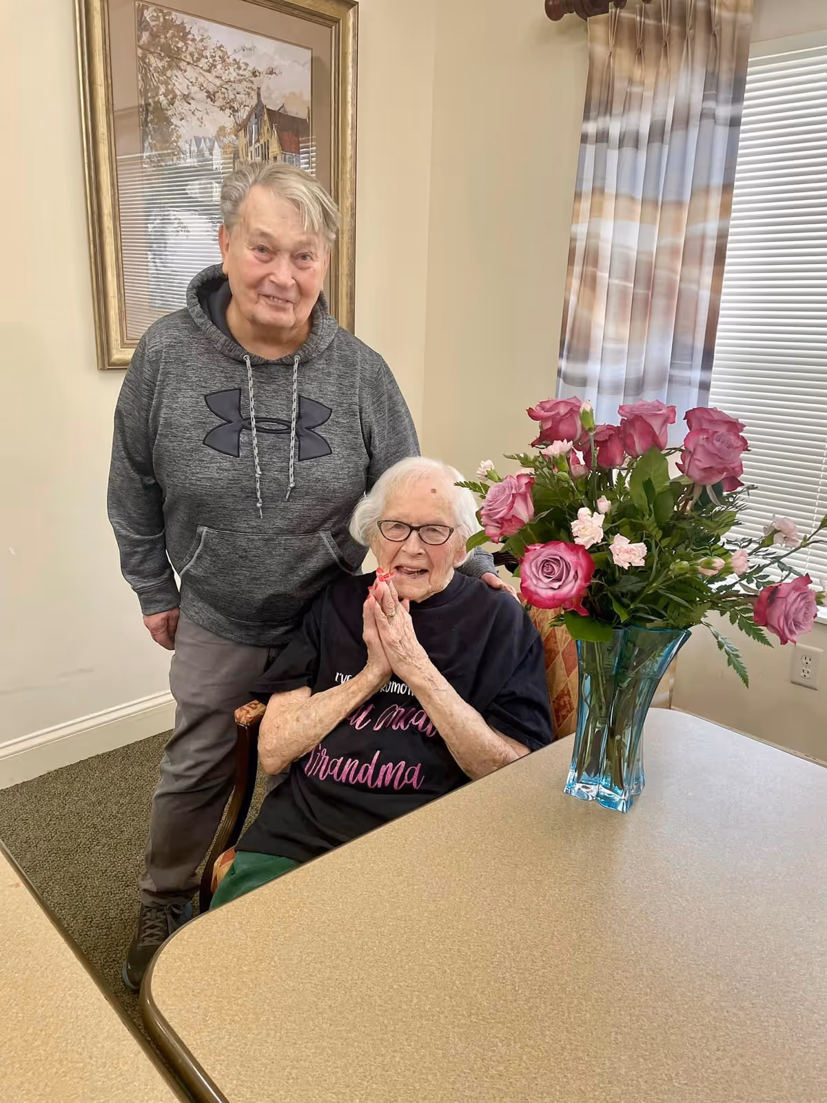 An elderly woman sitting at a table with a bouquet of pink roses in a blue vase, smiling and wearing glasses and a black shirt that says 'World's Greatest Grandma.' A man stands behind her, smiling and wearing a gray hoodie. The room has beige walls, a framed painting, and a window with striped curtains.