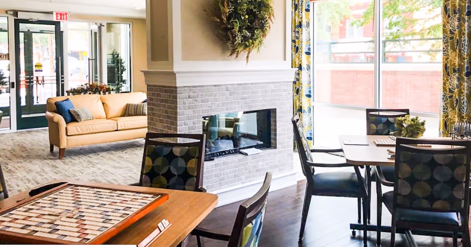 A bright and cozy common area in a senior living facility featuring a central fireplace with a wreath above it, several tables with chairs around them, one table with a board game, a beige sofa with cushions near the entrance, large windows with floral curtains letting in natural light, and a glass door exit in the background.