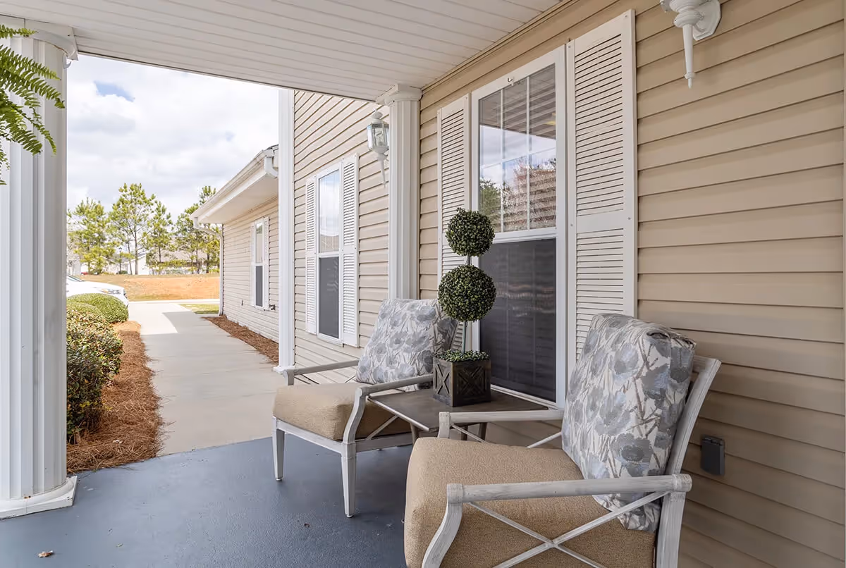 Covered porch area with two cushioned chairs and a small table with a decorative plant, beige siding with white shutters on the windows, and a concrete walkway leading away from the porch.