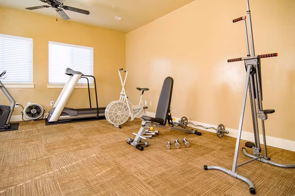 A small fitness room with exercise equipment including a treadmill, stationary bike, weight bench with dumbbells, and a multi-function workout machine. The room has beige walls, carpeted floor, two windows with blinds, and a ceiling fan.