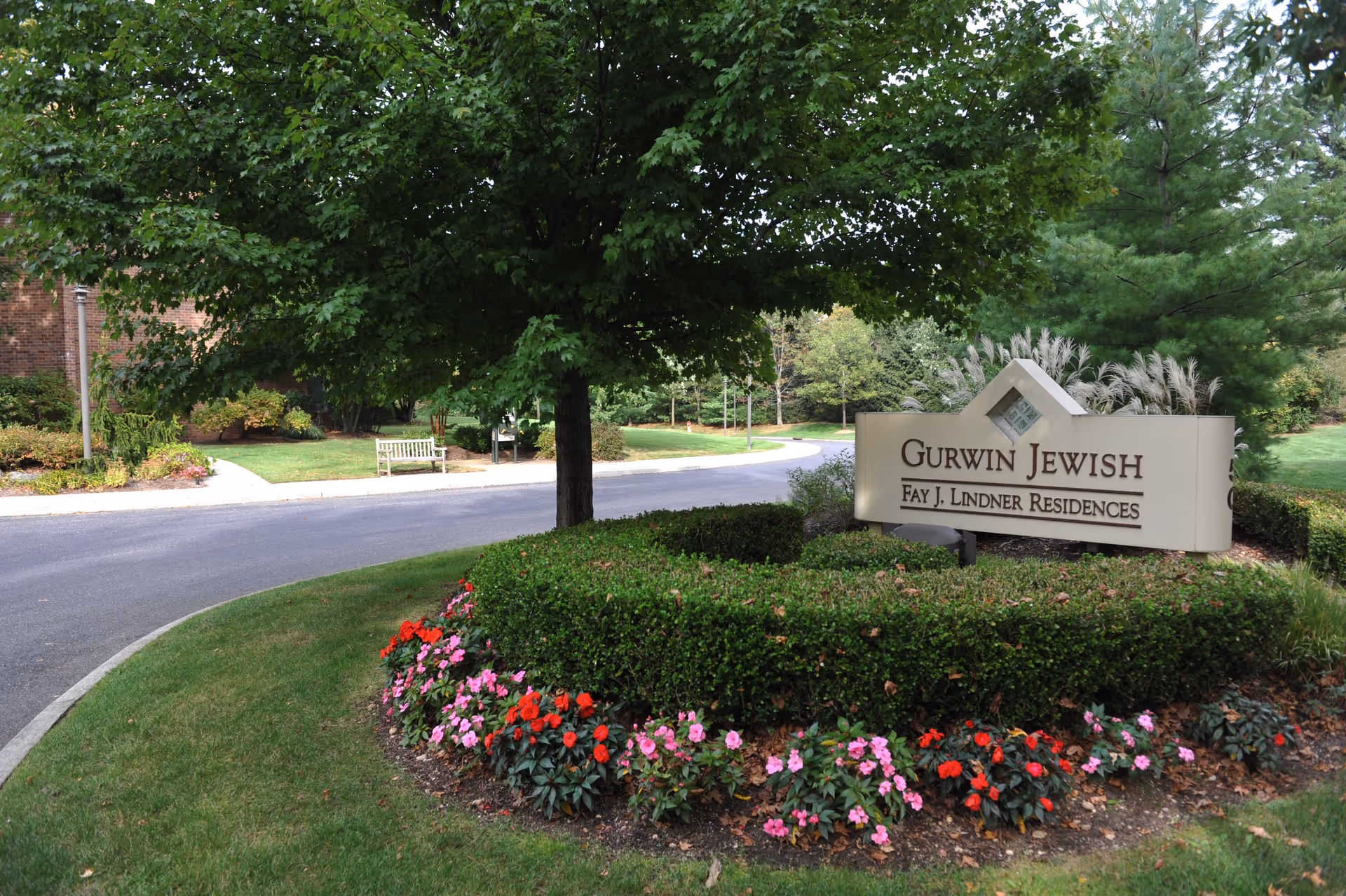 Outdoor view of the entrance area to Gurwin Jewish Fay J. Lindner Residences, featuring a landscaped garden with green bushes and colorful flowers surrounding a sign with the facility's name, a tree providing shade, a curved driveway, and a bench in the background.