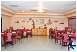 A dining room with multiple tables covered with red tablecloths and surrounded by chairs. The room has a ceiling fan with lights, a wooden dresser against the far wall decorated with a lamp, flowers, and wall art. The walls are light-colored with small shelves and decorative items.