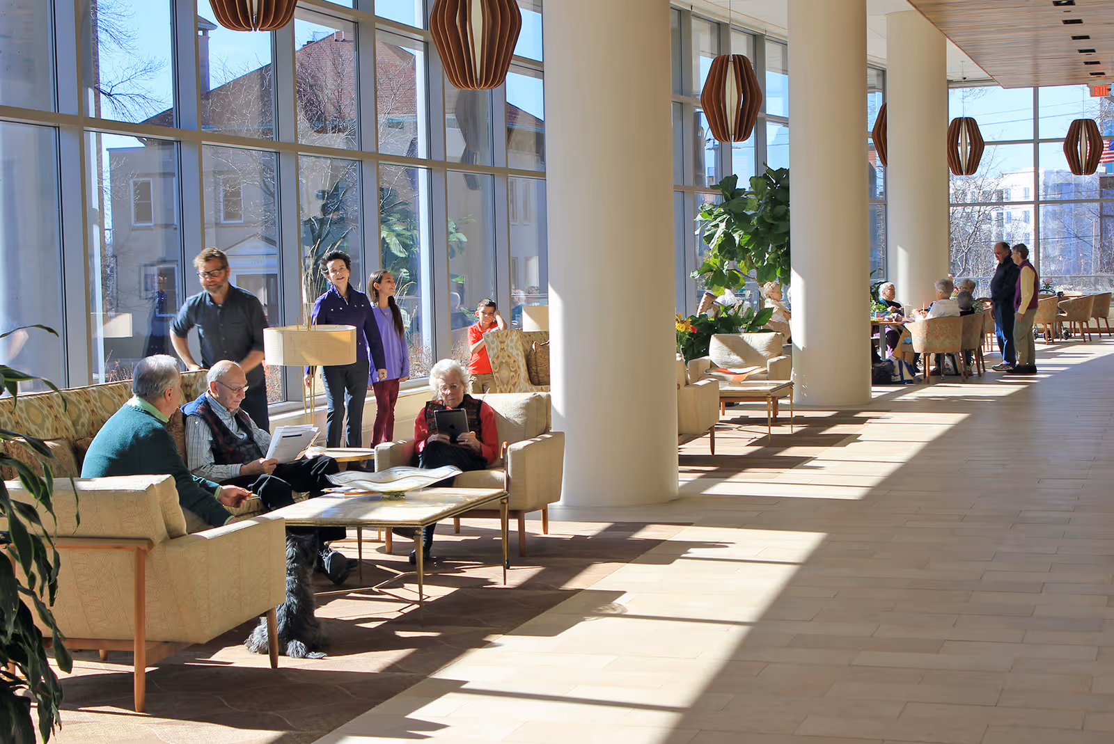 Sunlit senior living facility lounge with tall windows, columns, seating areas and residents socializing.