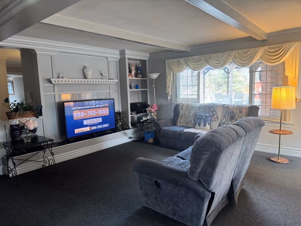 A cozy living room with two gray recliner sofas, a large window with cream-colored drapes, a standing lamp, and a flat-screen TV mounted on a white decorative wall. There is a small black table with flowers on the left and built-in shelves with decorative items on the right.