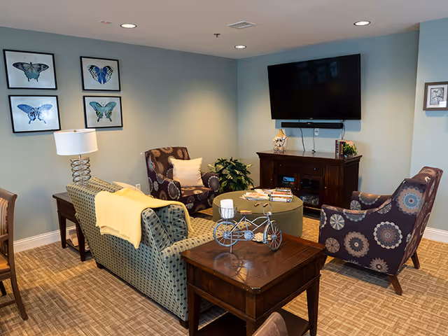 Cozy common living room with patterned chairs and a sofa arranged around a coffee table and wall-mounted TV, with butterfly artwork on the wall.