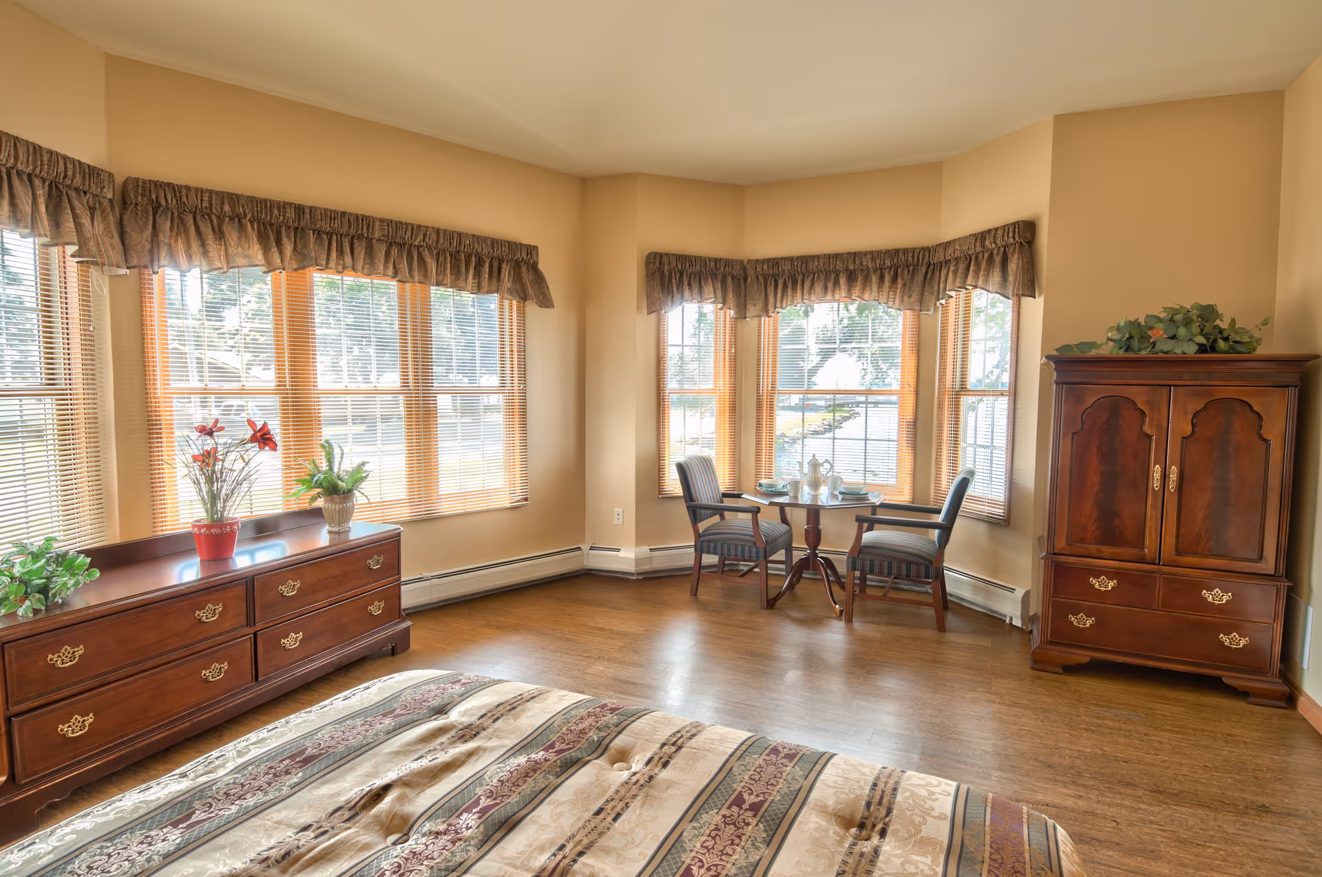 A bright bedroom with large windows covered by blinds and valances, featuring a wooden dresser with plants and flowers on top, a small round table set for two with chairs near the windows, and a wooden armoire. The bed with a patterned bedspread is partially visible in the foreground.