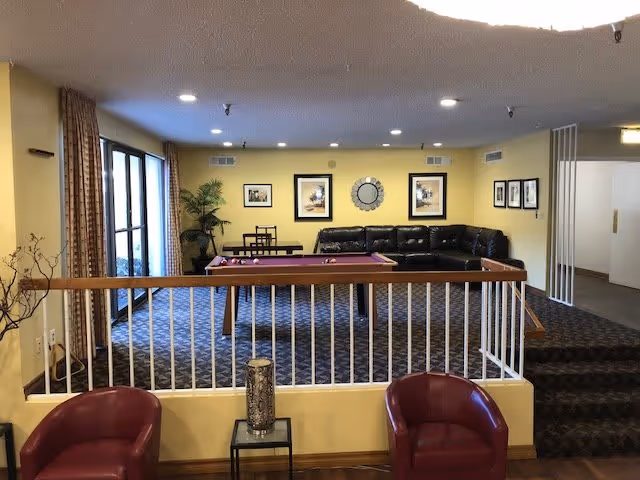 Interior view of a common area in a senior living facility with a pool table in the center, a black leather sectional sofa against a yellow wall with framed pictures and a round mirror, two red chairs in the foreground, a small table with a decorative vase, and large windows with curtains on the left side.