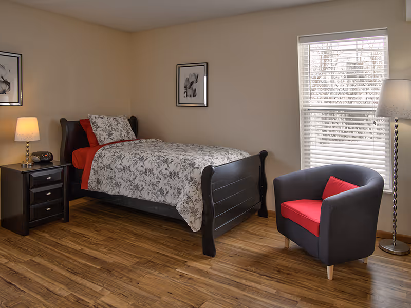 A cozy bedroom with a single dark wooden bed featuring floral bedding and red pillows. Next to the bed is a black nightstand with a lamp and an alarm clock. A dark armchair with a red cushion is positioned near a window with white blinds, and a tall floor lamp stands beside it. The room has wooden flooring and beige walls adorned with two framed black and white floral pictures.