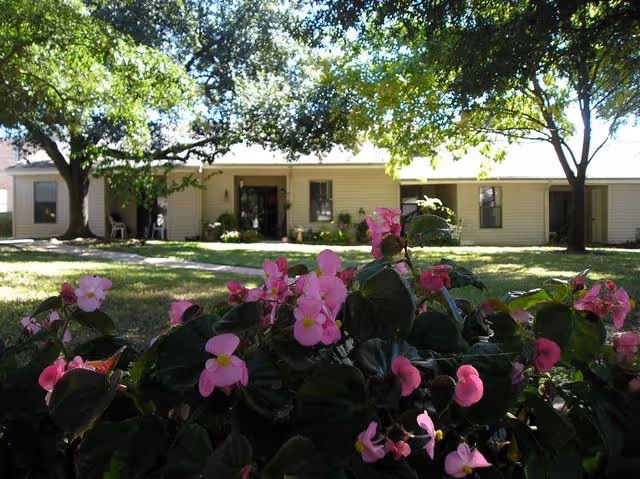 Pink flowers in the foreground with a shaded lawn and a single-story retirement community building under trees in the background.