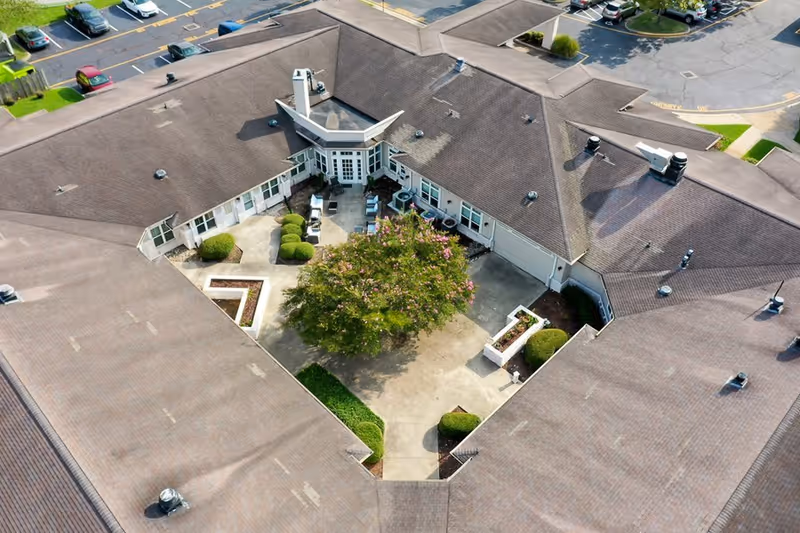 Aerial view of a senior living facility courtyard with a large tree in the center, surrounded by a paved area with shrubs and outdoor seating. The building has a brown roof and windows facing the courtyard. Parking spaces and cars are visible around the building.