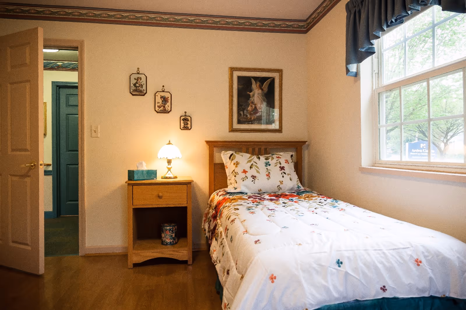 A cozy bedroom with a single bed covered in a white comforter with colorful floral patterns. Next to the bed is a wooden nightstand with a lit lamp and a box of tissues. The walls are decorated with small framed pictures and a larger framed artwork above the bed. A window with blue curtains lets in natural light, and an open door reveals a hallway with teal-colored doors.