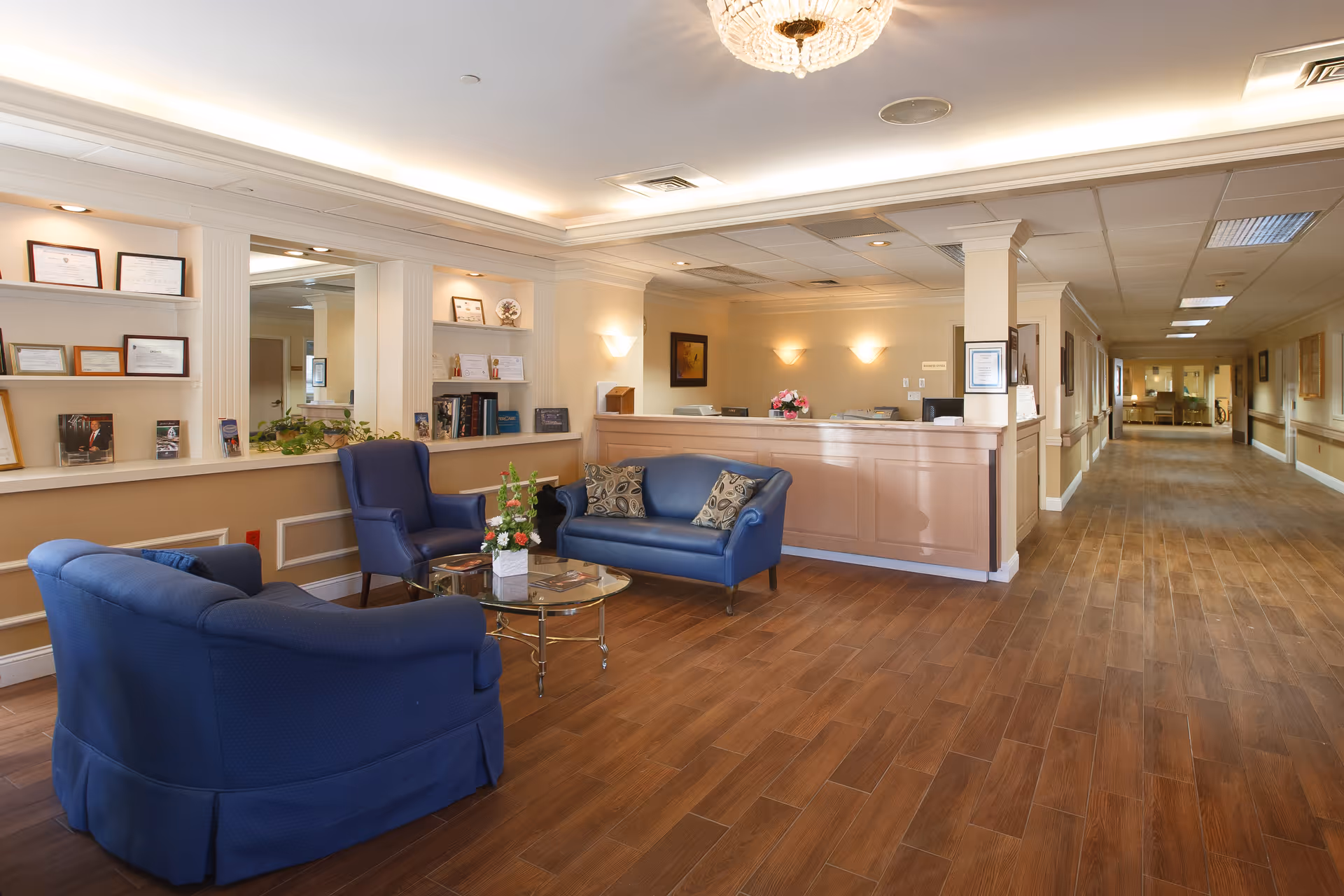 Reception lobby with blue sofas and chairs around a glass coffee table, a reception desk, and a long hallway.