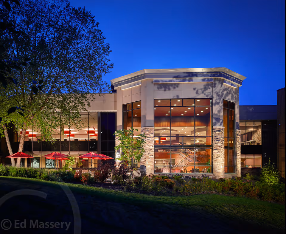 Exterior view of a modern building at dusk with large glass windows showing an illuminated interior with a staircase. The building is surrounded by landscaping with trees, bushes, and red patio umbrellas.