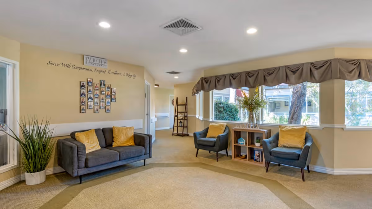 A bright and spacious living room area in a senior living facility with beige walls and carpet. The room features a gray sofa with yellow pillows, two blue armchairs each with a yellow pillow, a wooden shelving unit with decorative items and flowers in a vase, and large windows with brown valances letting in natural light. On the wall above the sofa is a collage of framed photos and a sign that reads 'Serve With Compassion, Respect, Excellence, & Integrity.'