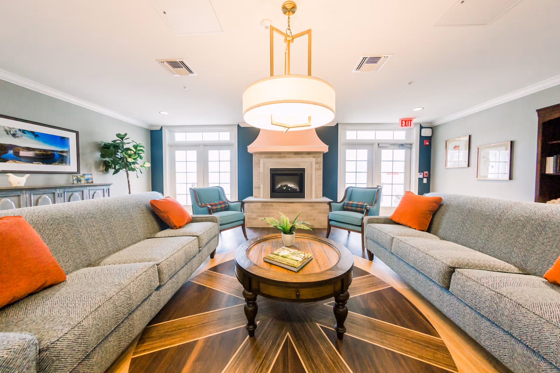 A bright and inviting senior living facility common area with two gray sofas adorned with orange pillows facing each other, a round wooden coffee table with a small plant and books, two teal armchairs with plaid cushions near a fireplace, large windows and glass doors letting in natural light, a modern ceiling light fixture, and framed artwork on the walls.