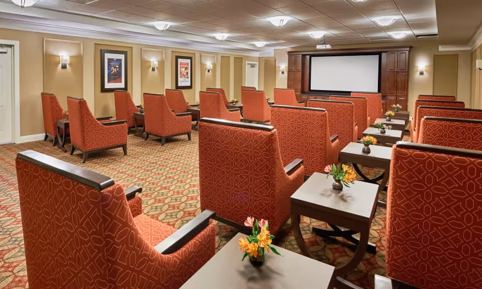 A cozy theater room with multiple rows of red patterned armchairs facing a large blank projection screen. Each chair has a small side table with a small flower arrangement. The room has beige walls with framed artwork and soft wall lighting, and a patterned carpet floor.