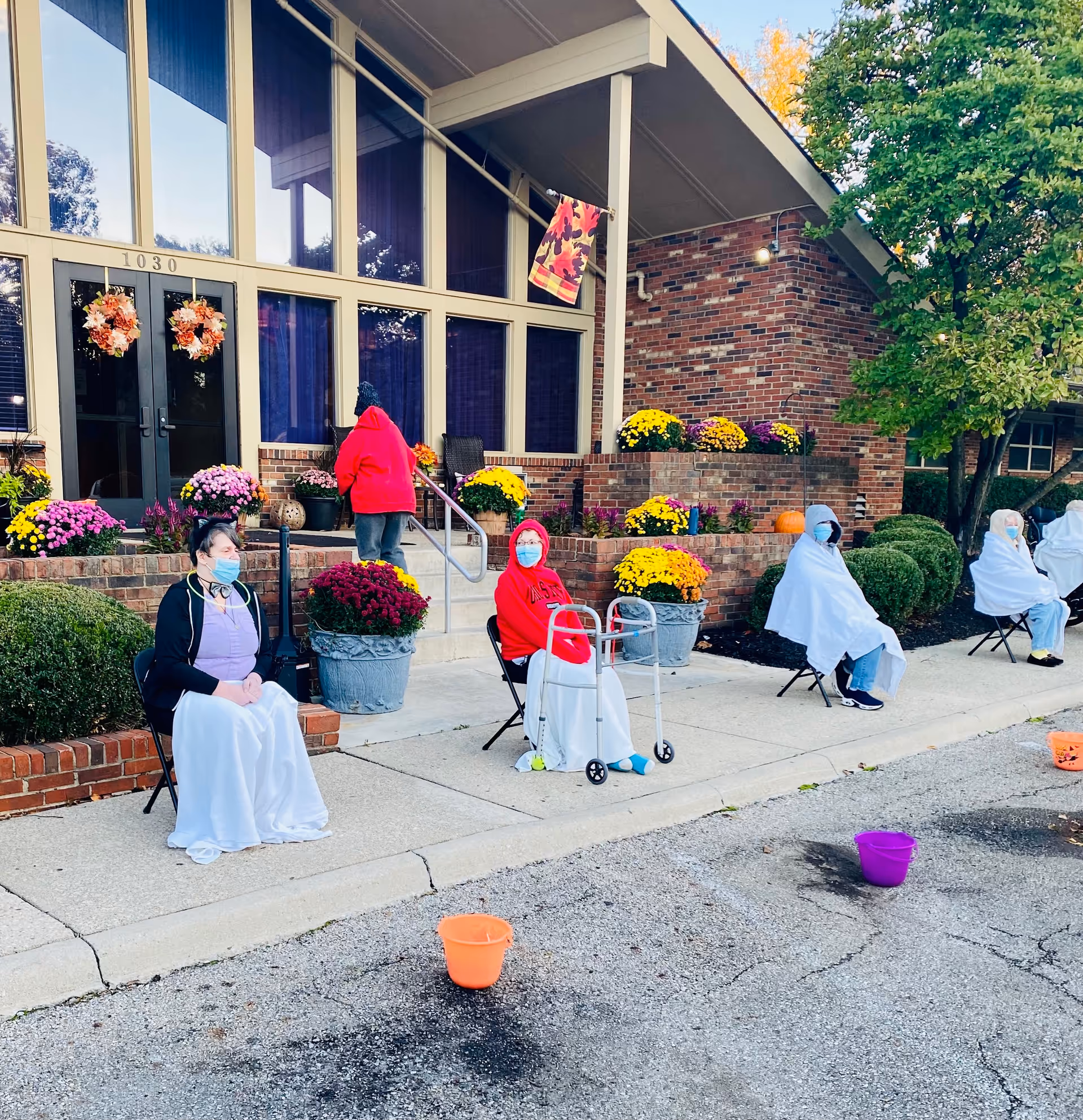 Several elderly individuals wearing masks and wrapped in white blankets are seated on chairs spaced apart on a sidewalk outside a brick building decorated with colorful flowers and autumn wreaths. One person in a red hoodie is using a walker, and another person in a red jacket is walking up the steps toward the building entrance. Orange and purple buckets are placed on the street in front of them.