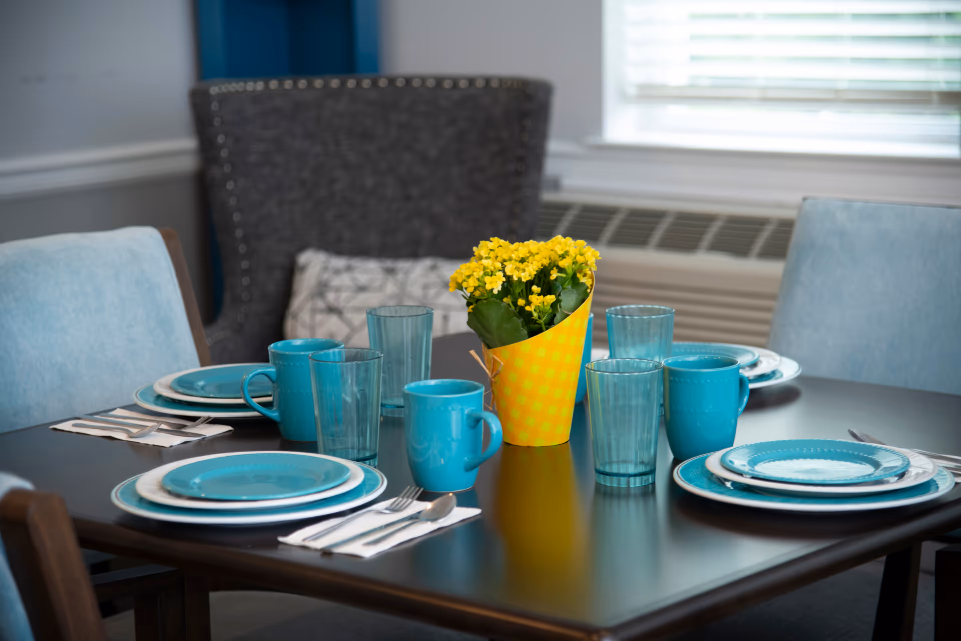 A dining table set for four with turquoise plates, mugs, and glasses. A small yellow potted plant is placed in the center of the table. The background shows a cushioned chair and a window with blinds.