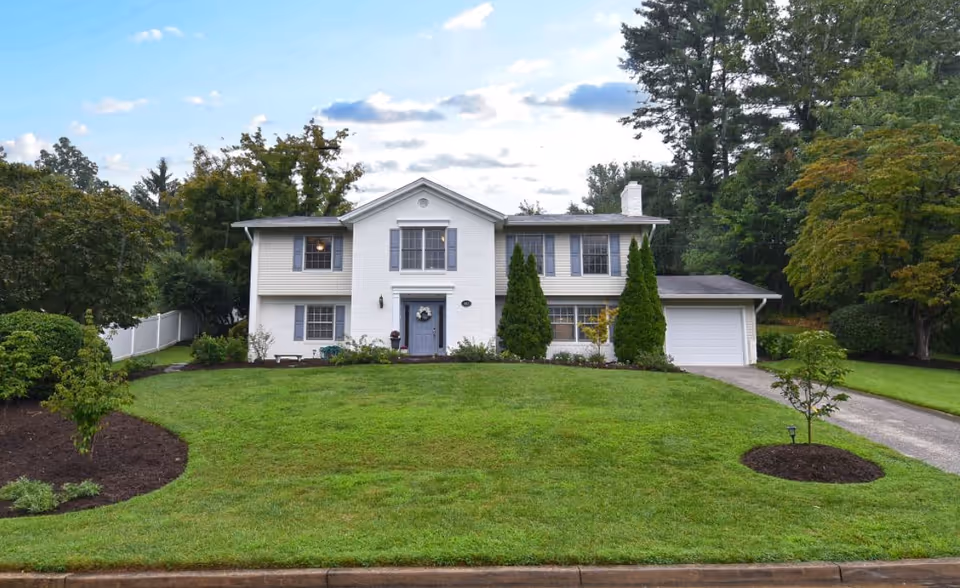 Front exterior view of a two-story white house with a well-maintained green lawn, trees, and shrubs under a partly cloudy sky.