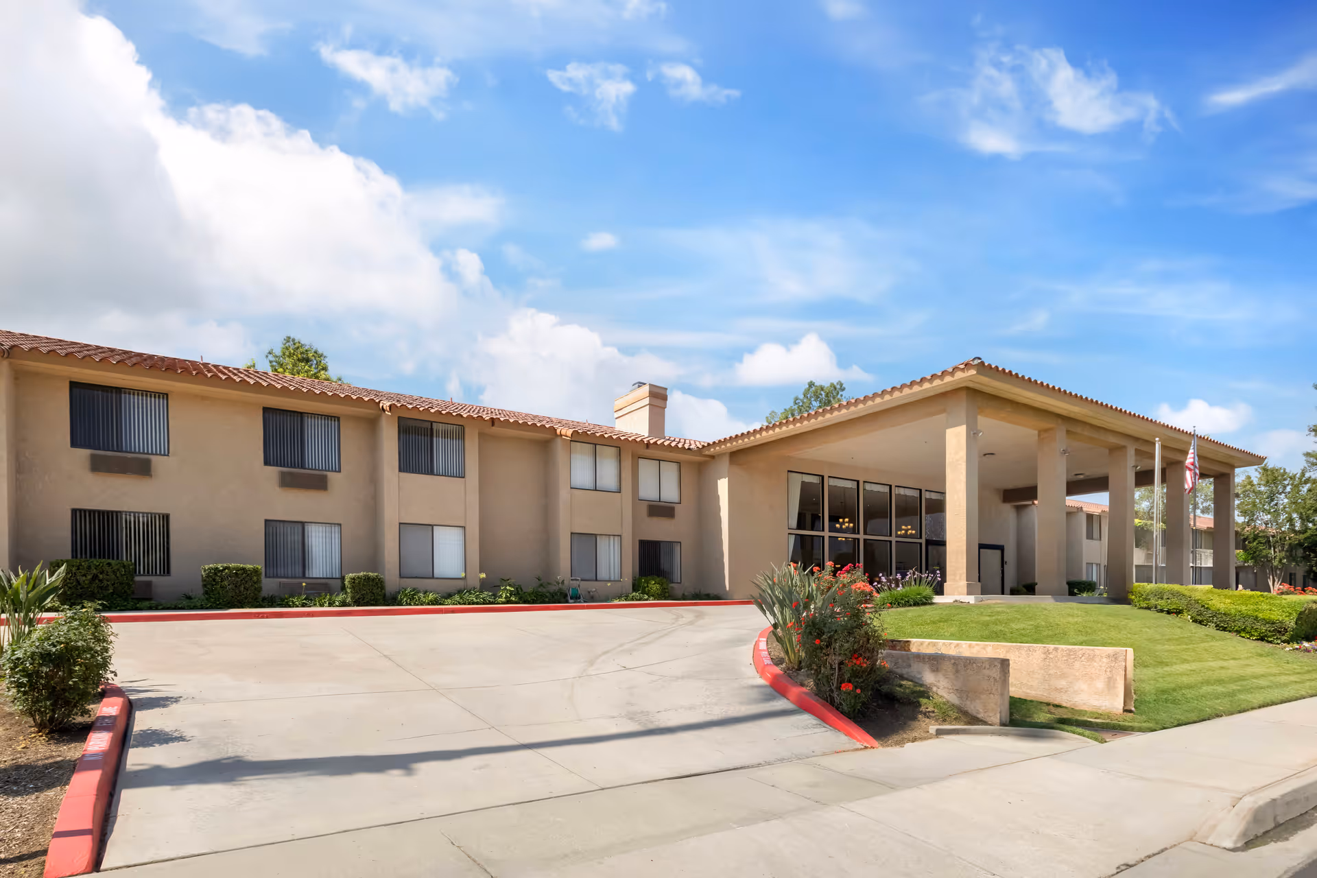 Exterior view of a two-story senior living facility building with beige stucco walls and a red tile roof under a partly cloudy blue sky. The entrance has a covered driveway with large pillars, landscaped greenery, and an American flag on a flagpole.