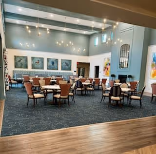 A spacious dining room with multiple round tables and chairs arranged neatly. The room has high ceilings with modern light fixtures and large windows allowing natural light. The walls are decorated with framed artwork and a large mirror, and the floor has a combination of carpet and wood.