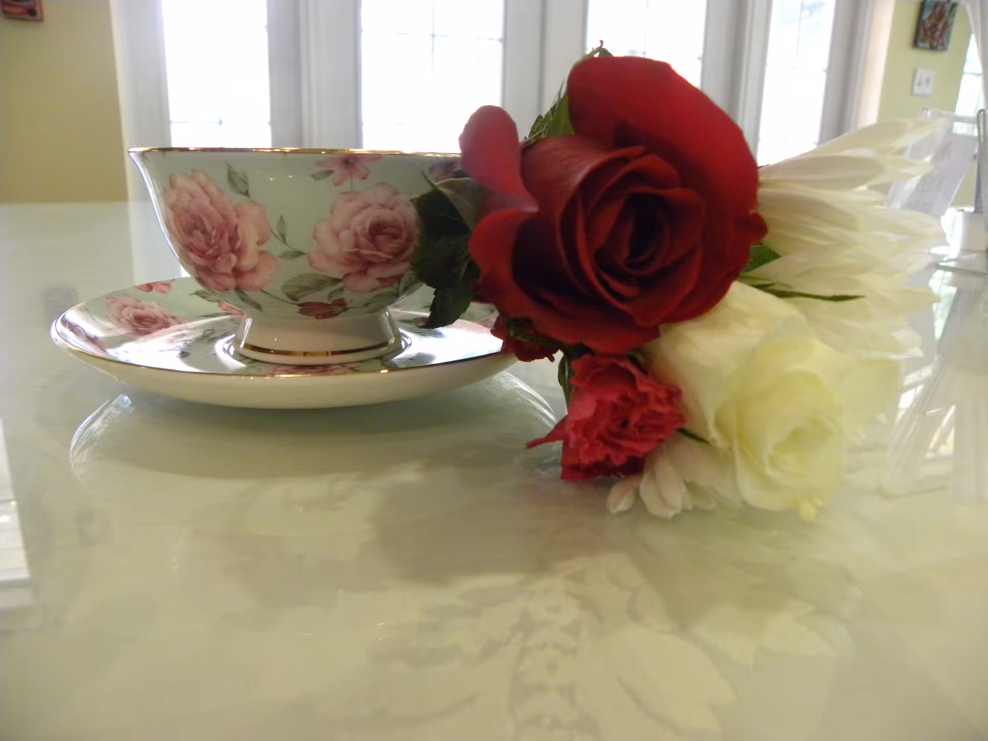 A floral teacup and saucer beside a small bouquet of red and white roses on a glossy table.