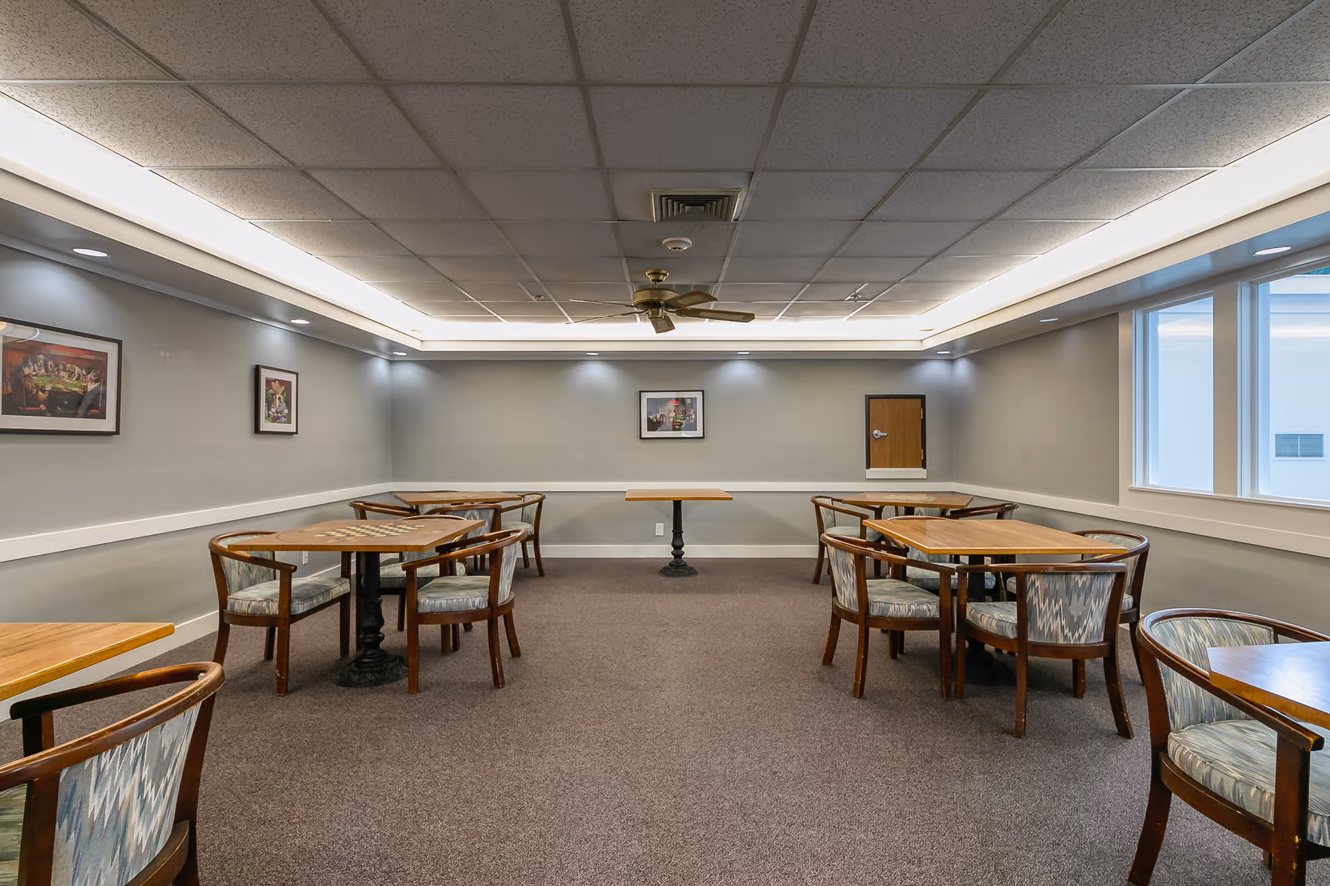 A well-lit room with several wooden tables and cushioned chairs arranged neatly. The walls are painted gray with white trim, and there are framed pictures hanging on the walls. The ceiling has recessed lighting and a ceiling fan in the center. Large windows on the right side allow natural light to enter the room.