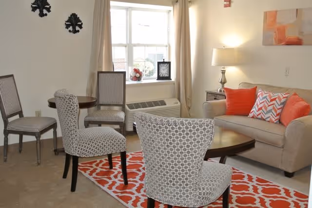 A cozy living room with a beige sofa adorned with orange and patterned throw pillows, a wooden coffee table, and four chairs with different upholstery patterns arranged around the table. The room has a window with beige curtains, a table lamp on a side table, and decorative wall art. A red and white patterned rug is on the floor.