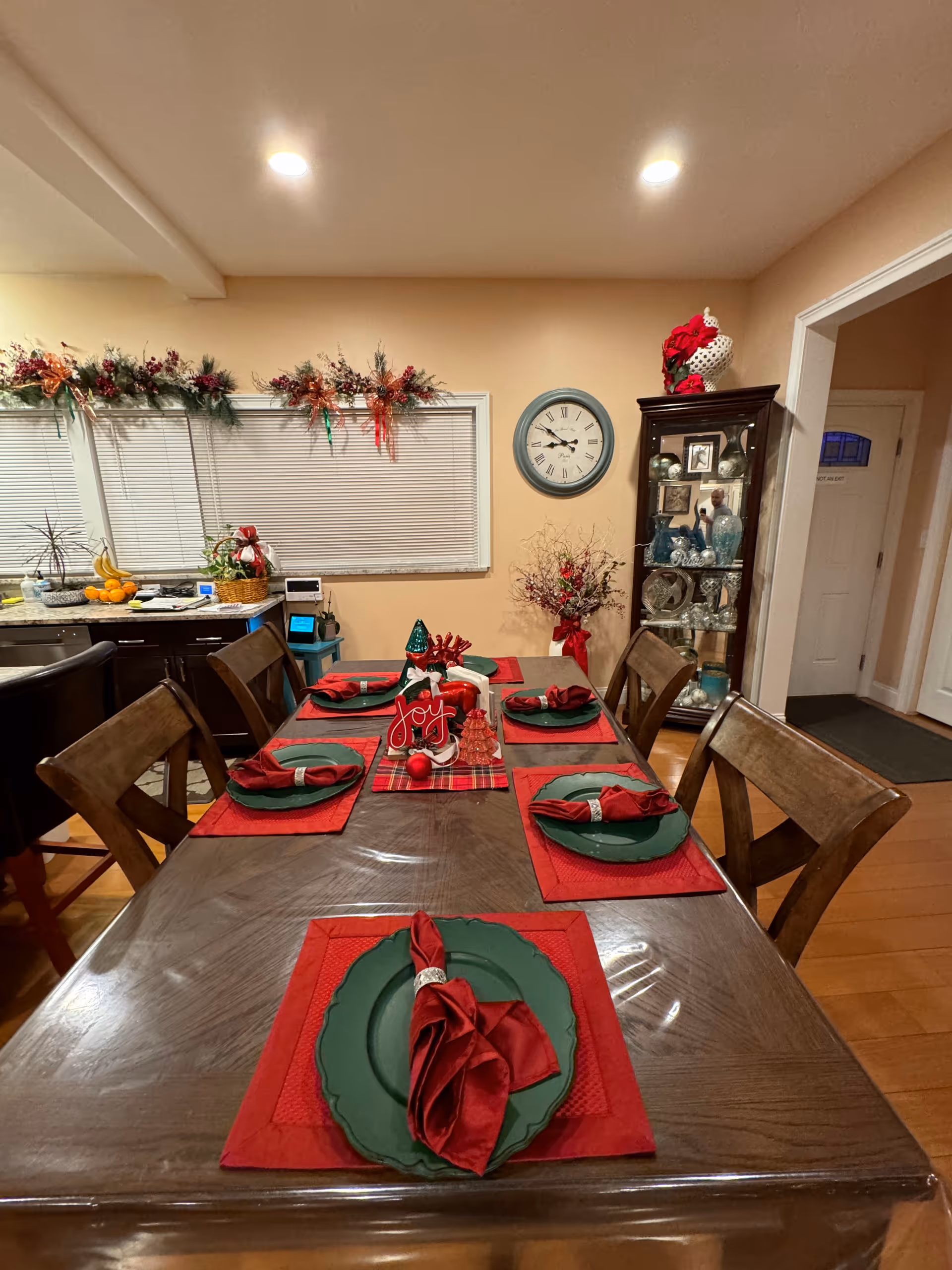 Dining room with a long wooden table set for a holiday meal with red and green place settings, window garlands, and a clock and china cabinet on the wall.