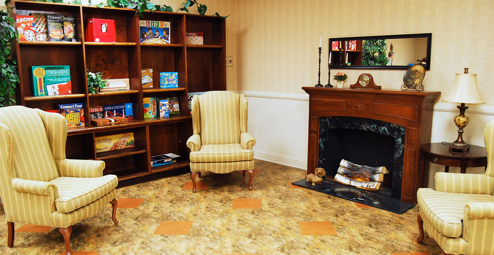 A cozy living room area with three striped armchairs arranged around a wooden fireplace with a black marble surround. The fireplace mantle holds decorative items including a clock, candlesticks, a small plant, and a decorative plate. Behind one armchair is a wooden bookshelf filled with various board games. A table with a lamp is positioned next to one of the chairs. The room has a warm, inviting atmosphere with patterned flooring and light-colored wallpaper.