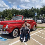 A person in a wheelchair and another person standing behind them smiling in a parking lot next to a vintage red convertible car. There are trees and other parked cars in the background under a partly cloudy sky.