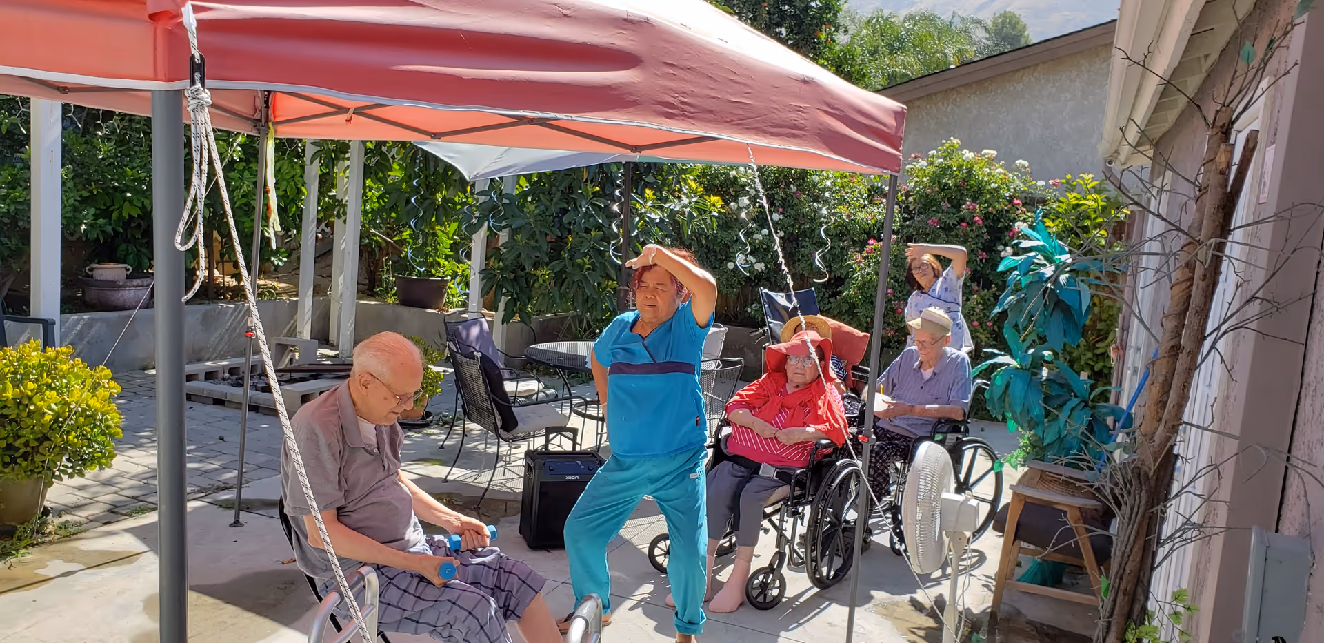 An outdoor patio area at Terrace Gardens with elderly residents and a caregiver. One elderly man is seated and lifting small dumbbells, a caregiver in blue scrubs is standing and stretching, and two elderly women in wheelchairs are seated nearby. The area is shaded by a red canopy with greenery and flowers in the background.