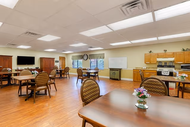 Bright communal dining and activity room with multiple tables and chairs, a kitchenette, TV, and wood floors.