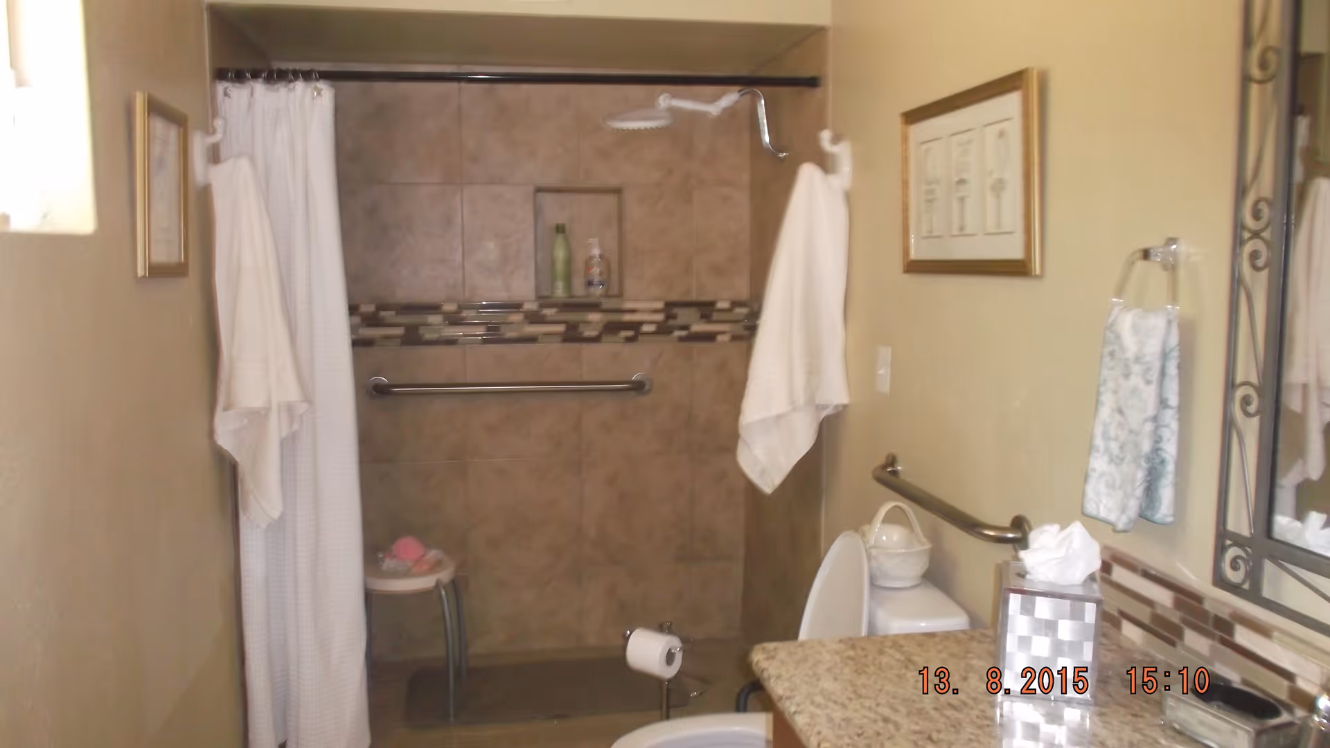 A bathroom with a walk-in shower featuring beige tiles and a decorative tile border. The shower has a white curtain, a wall-mounted showerhead, and a grab bar. There is a small stool inside the shower with bath items on it. A white towel hangs on a hook outside the shower. The bathroom also includes a toilet with a grab bar beside it, a granite countertop with a tissue box, and a framed picture on the wall.