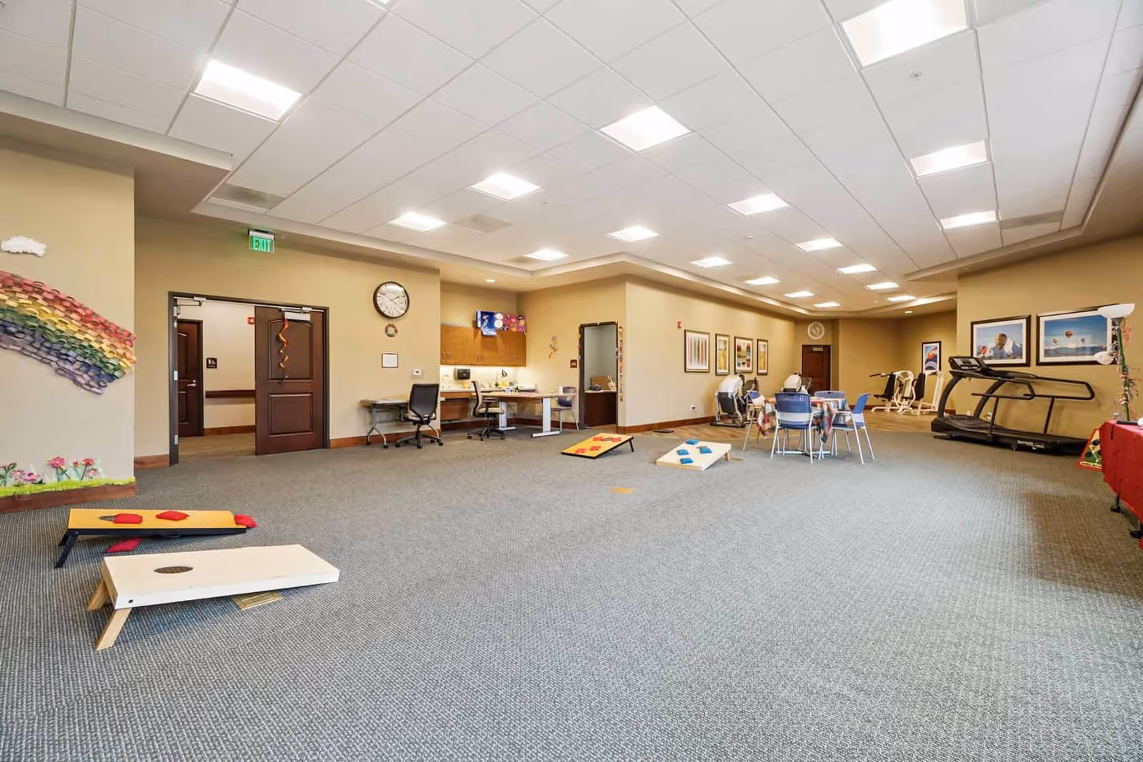 Spacious multi-purpose activity room with cornhole boards on the floor, tables and chairs, exercise equipment, and a reception/desk area.
