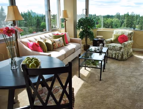 Bright, furnished living room with a sofa, patterned armchair, glass coffee tables, round dining table with apples, and large windows overlooking a wooded view.