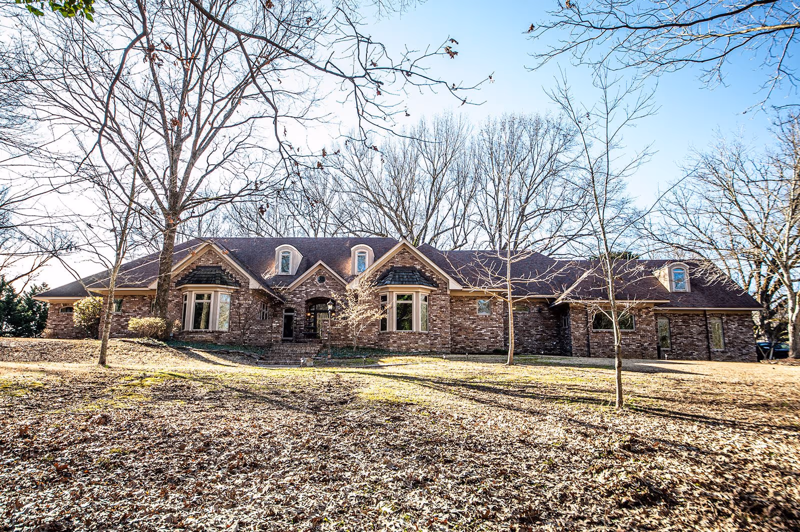 Front view of a large brick single-story building with dormer windows surrounded by leafless trees on a sunlit lot.