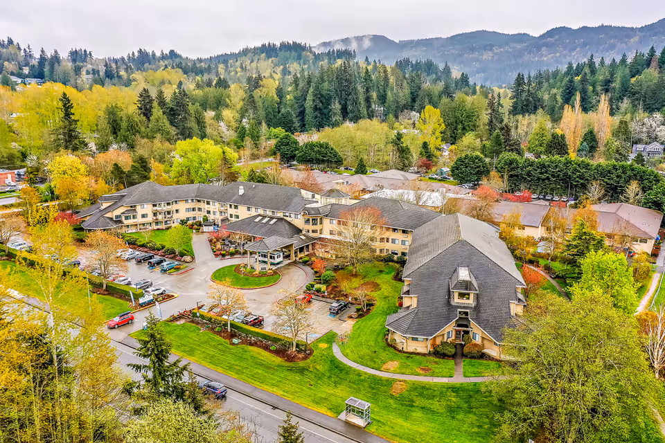 Aerial view of Solstice Senior Living at Bellingham, showing a large senior living facility surrounded by lush greenery and trees with a mountainous backdrop. The building has multiple wings with a central entrance and parking areas with cars parked. The landscape includes well-maintained lawns and pathways.
